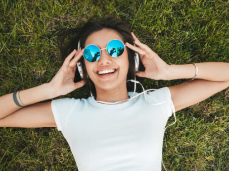 Fashion portrait of young stylish hipster woman lying on the grass in the park .Girl weares trendy outfit.Smiling model enjoy her weekends. Female listening to music via headphones.Top view casque de musique